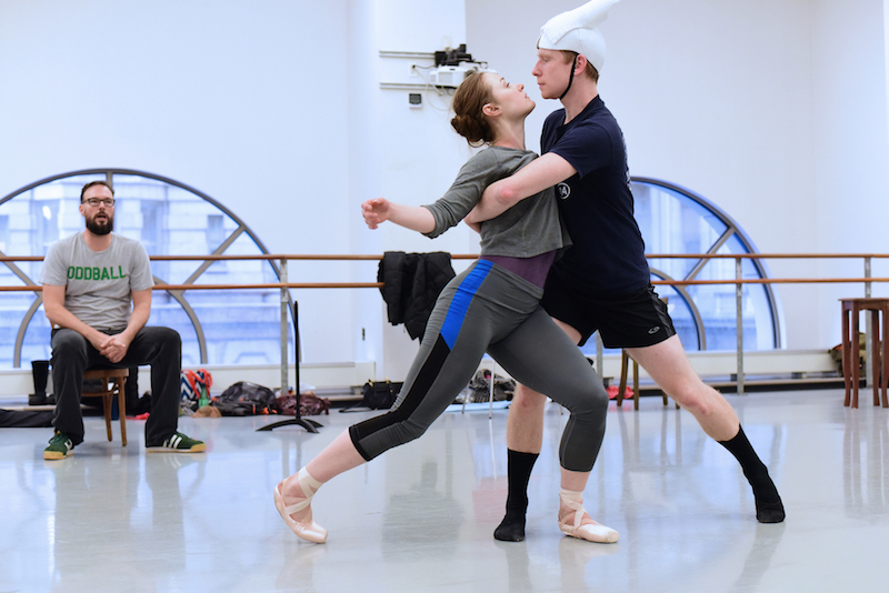 Choreographer Trey McIntyre wearing sneakers, jeans and glass sits at the back of a windowed studio while two dancers, one in pointe shoes and other wearing a pointed white headpiece, are locked in an embrace